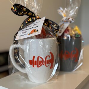 May include: Two wrapped mugs, one white and one gray, are displayed. The white mug has a red graphic and the letters "LDC". The gray mug has a similar red graphic. Each mug is wrapped in clear cellophane and tied with a decorative ribbon.