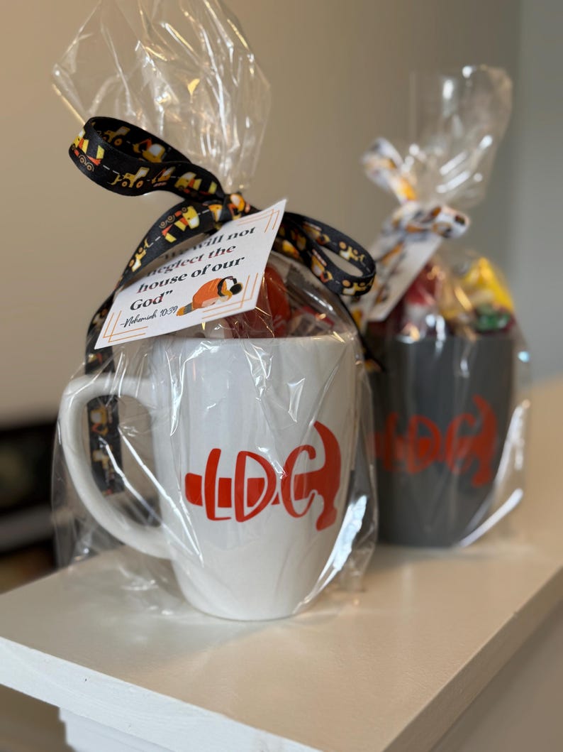 May include: Two wrapped mugs, one white and one gray, are displayed. The white mug has a red graphic and the letters "LDC". The gray mug has a similar red graphic. Each mug is wrapped in clear cellophane and tied with a decorative ribbon.
