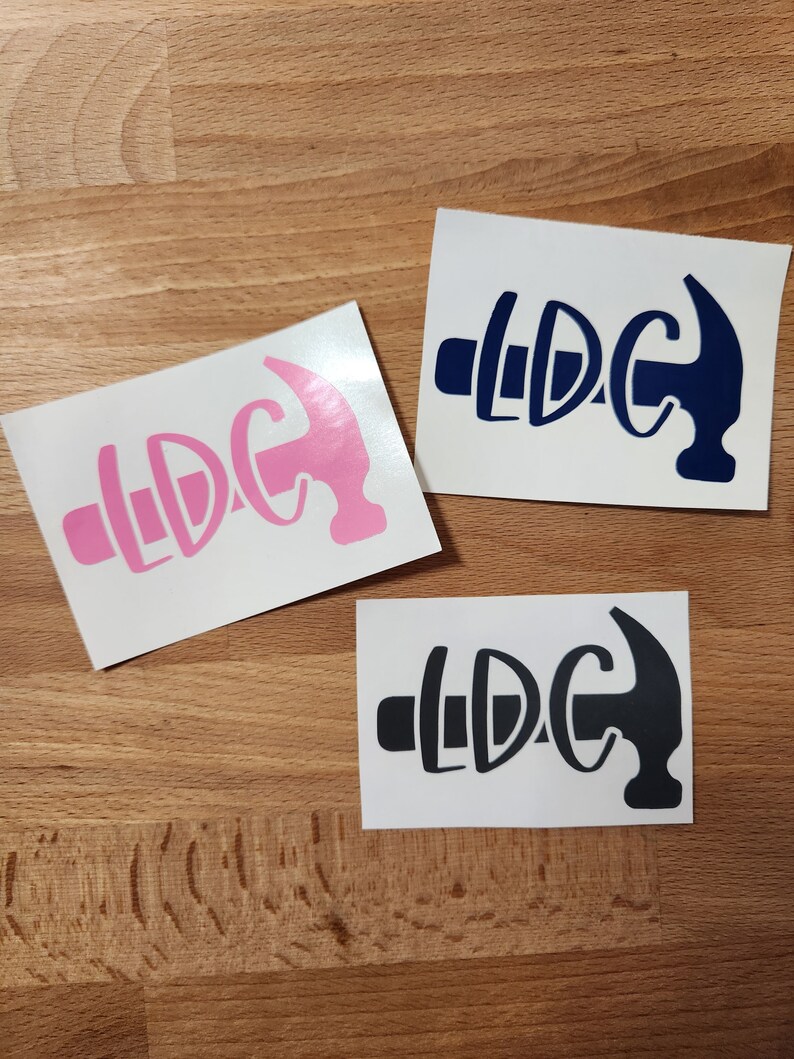 May include: Three vinyl stickers with a hammer design and the letters "LDC". One sticker is pink, one is navy blue, and one is black. The stickers are on a light brown wooden surface.