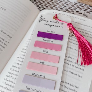May include: A white rectangular bookmark featuring the text my reading companion and a pink tassel. It includes six adhesive tabs in shades of purple, pink, and lavender, each labeled with phrases like quotes, favorites, omg, wtf, plot twist, and i'm not okay.