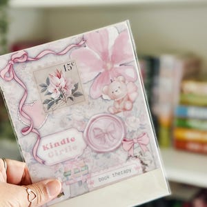 May include: A clear plastic sleeve containing stickers with a floral and teddy bear theme. The stickers feature pink and white colors, with text that reads "Kindle Girlie" and "book therapy". A postage stamp with a floral design is also visible.