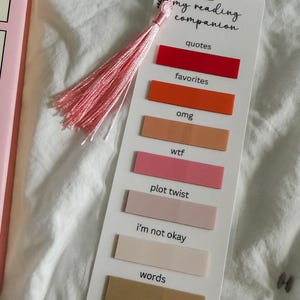 May include: A white rectangular bookmark featuring a pink tassel and the text "my reading companion" at the top. It includes six horizontal adhesive tabs in shades of red, orange, peach, pink, light mauve, beige, and tan, labeled with text.