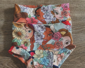Puede incluir: Un par de bombachos de bebé blancos con un estampado colorido que presenta princesas de Disney rodeadas de flores. Las princesas incluyen Pocahontas, Bella, Ariel y Cenicienta.