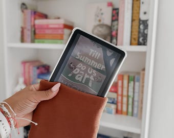 May include: A rust-colored tablet sleeve holding a tablet with the text "Till Summer Do Us Part". The sleeve is made of a soft, textured material. A white bookshelf filled with books is in the background.