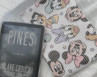 Pode incluir: Um leitor de e-books Kindle com o livro "Pines" de Blake Crouch, ao lado de um tecido branco com um padrão de personagens de desenhos animados, incluindo Mickey e Minnie Mouse, Pato Donald e Pateta. O leitor está em uma capa transparente.