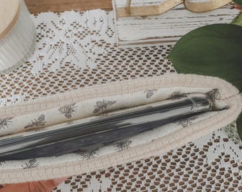 May include: A beige utensil holder with a bee pattern, holding two silver utensils. The holder is made of a soft fabric and is resting on a white lace tablecloth. A gold-colored decorative item is in the background.
