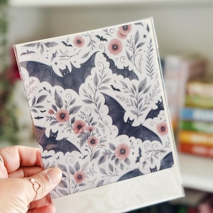 May include: A greeting card with a floral and bat pattern. The design features dark gray bats, pink flowers, and gray leaves on a white background. The card is held in a clear plastic sleeve.