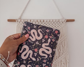 Könnte beinhalten: Eine dunkelblaue Clutch mit einem sich wiederholenden Muster aus rosa Schlangen, Blumen und Monden. Die Clutch wird in einer Hand gehalten, im Hintergrund befindet sich ein Makramee-Wandbehang.