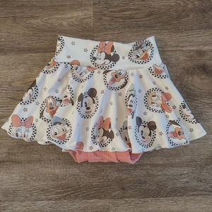 Puede incluir: Una falda blanca y rosa con volantes con un estampado de Mickey Mouse, Minnie Mouse, Donald Duck y Goofy. La falda tiene un fondo blanco con círculos a cuadros negros y blancos alrededor de cada personaje. La falda tiene una cintura elástica rosa.