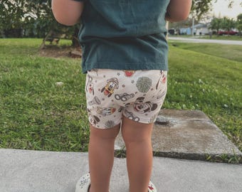 May include: A child wearing a teal t-shirt and white shorts with a Mickey Mouse and other Disney character print. The child is wearing white shoes with a Mickey Mouse design.
