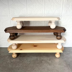 May include: A set of three tiered wooden display stands with turned wooden legs. The top tier is natural wood, the middle tier is stained brown, and the bottom tier is painted white.