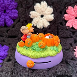 以下が含まれることがあります： 紫色の円形ハロウィーンをテーマにした置物。漫画のキャラクター、カボチャ、緑の草が描かれています。置物の前面にはスロットがあり、紫、白、ピンクのかぎ針編みの花に囲まれています。