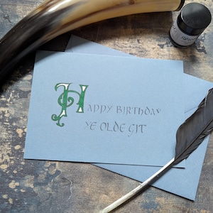 May include: A blue card with a green and white letter H and the text "Happy Birthday Ye Olde Git" written in calligraphy. A black feather quill pen and a bottle of ink are on the table.
