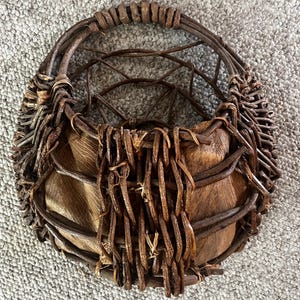 May include: A handcrafted, round basket made from natural brown twigs and woven materials. The basket features a sturdy handle and a textured, organic design, perfect for decorative storage or display.