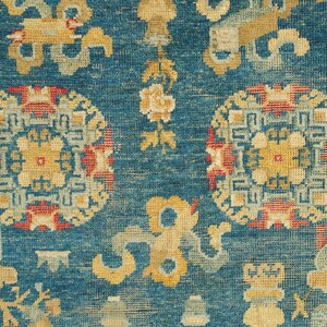 May include: A close-up of a blue rug with intricate patterns in shades of yellow, red, and white. The rug features repeating circular motifs with geometric designs and floral elements.