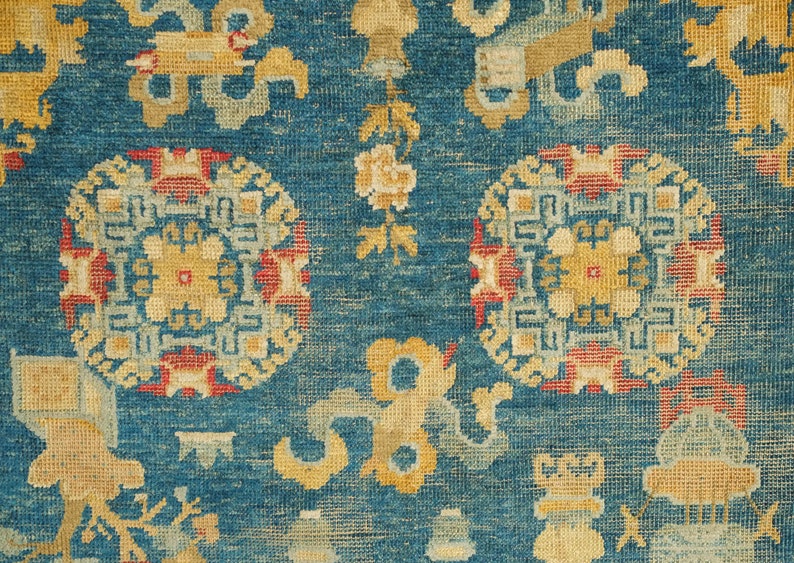 May include: A close-up of a blue rug with intricate patterns in shades of yellow, red, and white. The rug features repeating circular motifs with geometric designs and floral elements.