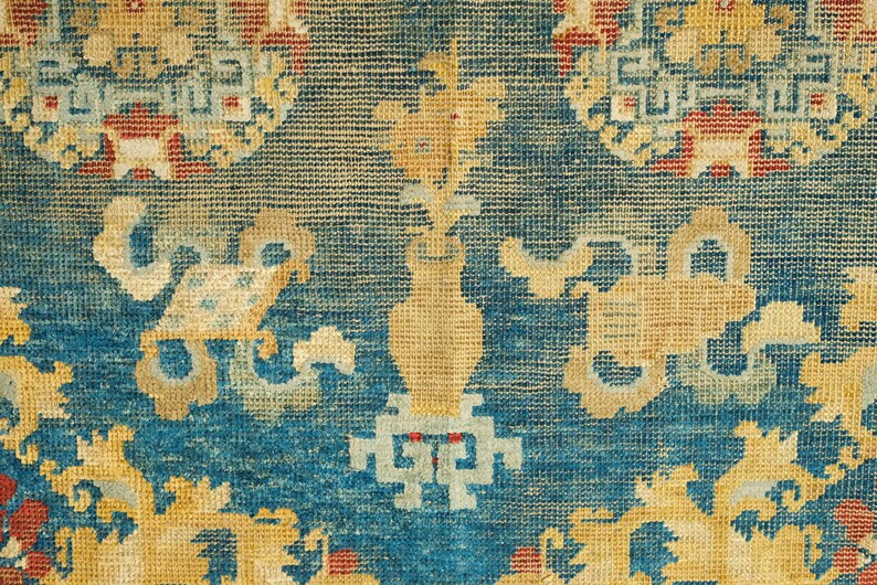May include: Close-up of a blue and yellow patterned rug with intricate designs. The rug has a faded, vintage look and features a repeating pattern of floral motifs.