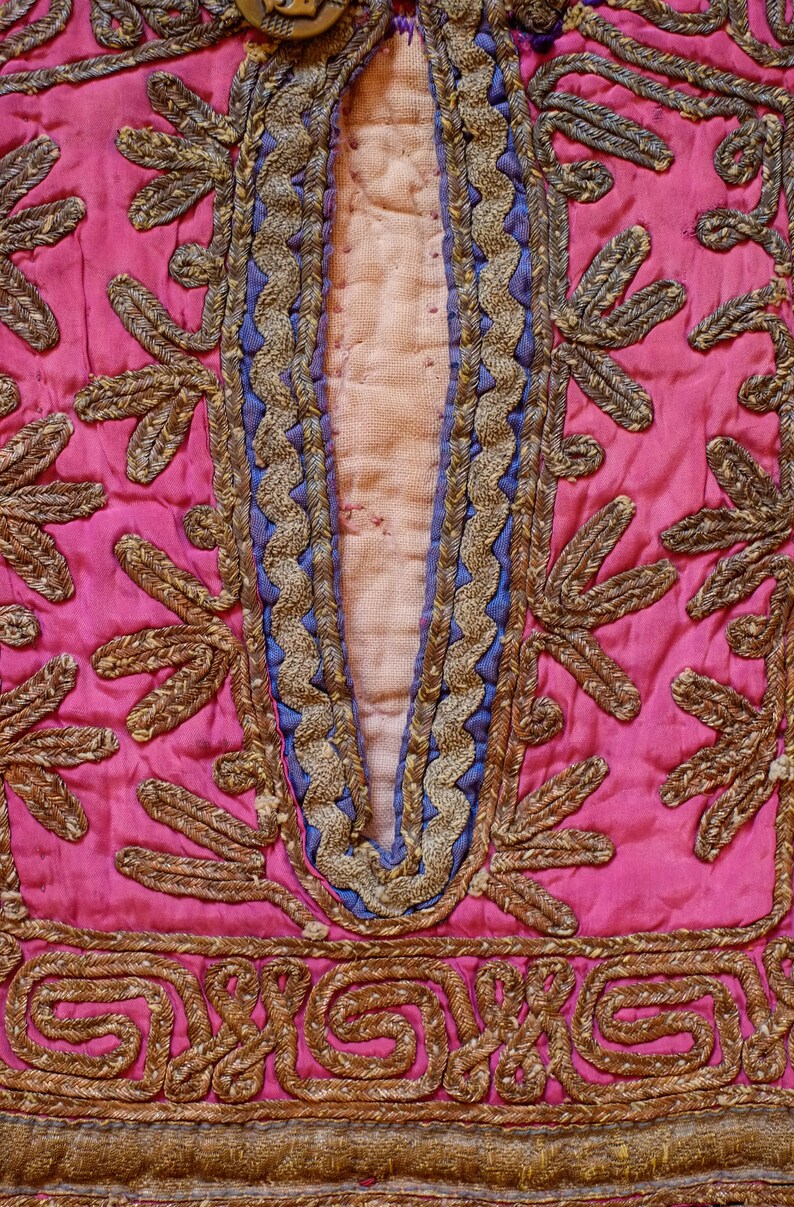 May include: Close-up of a pink fabric with intricate gold embroidery. The embroidery features a repeating pattern of leaves and swirls.