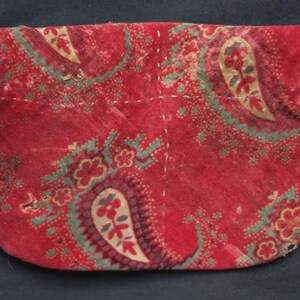 May include: A red fabric pouch with a paisley pattern. The pouch is sewn with white thread and has a rounded bottom.