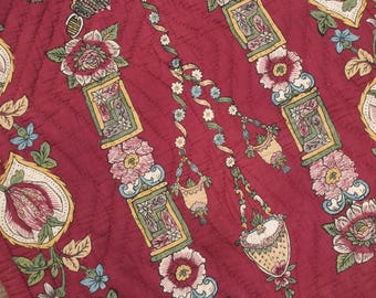 Puede incluir: Una tela roja acolchada con un patrón floral. El patrón presenta un diseño repetitivo de flores estilizadas, hojas y cestas colgantes. La tela tiene un aspecto y una sensación vintage.
