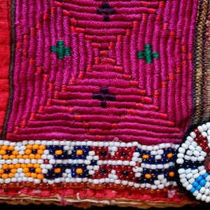 May include: A close-up of a colorful embroidered fabric with a beaded trim and a round beaded embellishment. The fabric is a mix of red, pink, white, and blue.