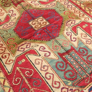 Può includere: Primo piano di un tappeto fantasia dal design tradizionale. Il tappeto presenta un campo centrale rosso con forme geometriche blu, verdi e crema. Il bordo presenta un motivo ripetuto in colori simili. La trama del tappeto è visibile.