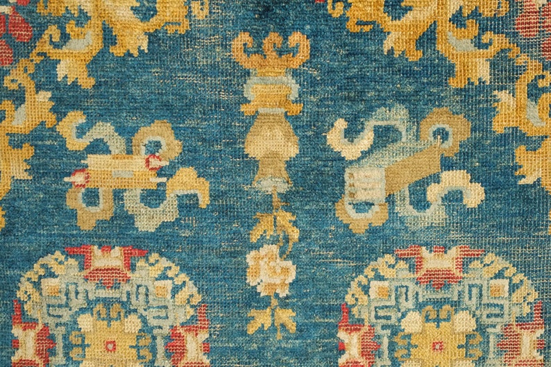 May include: Close-up of a blue rug with intricate patterns in shades of yellow, red, and white. The rug features a repeating design of stylized floral motifs and geometric shapes.
