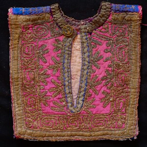 May include: A pink fabric garment with gold and blue embroidery. The garment has a decorative button and a slit down the front.