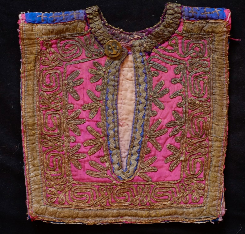 May include: A pink fabric garment with gold and blue embroidery. The garment has a decorative button and a slit down the front.