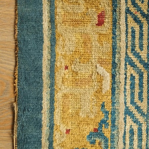 May include: Close-up of a blue and yellow rug with a geometric pattern. The rug has a faded appearance and shows signs of wear.