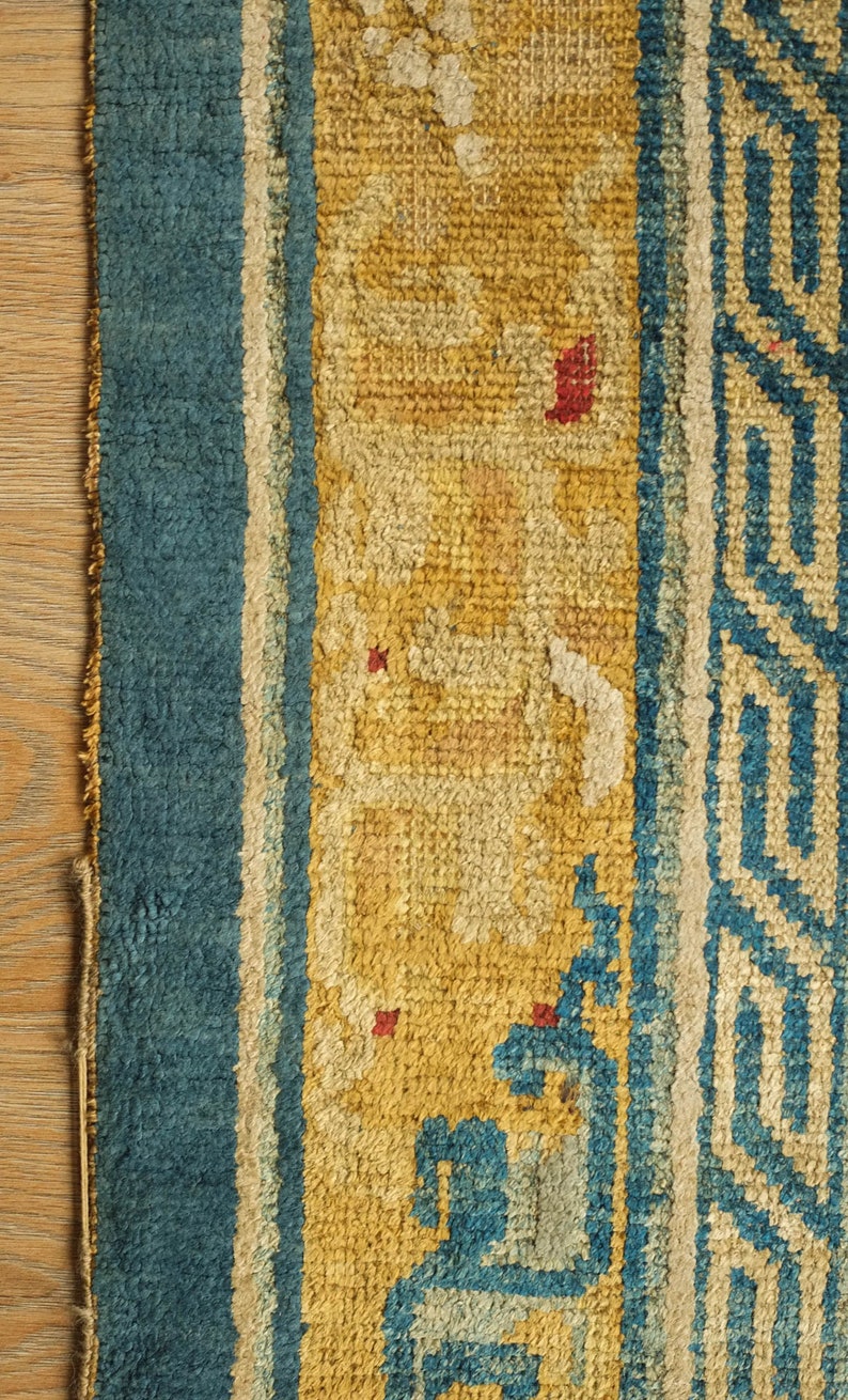 May include: Close-up of a blue and yellow rug with a geometric pattern. The rug has a faded appearance and shows signs of wear.