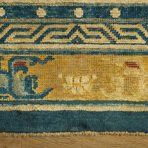 May include: Close-up of the bottom edge of a blue and yellow rug with a geometric pattern. The rug has a fringe border and is made of woven fabric.