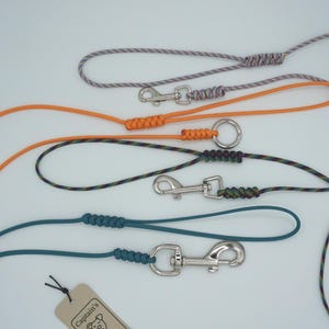 May include: Four dog leashes made of rope in various colors including orange, teal, and multi-colored patterns. Each leash features a silver metal swivel snap hook and a hand loop secured with a braided knot. A small tan tag with the text Captain's is visible.