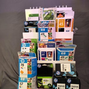 May include: A display of various HP inkjet printer ink cartridges in a white tiered shelf. Cartridges are in boxes with colors like yellow, orange, blue, and black. The shelf includes cartridges labeled 33, 45, 49, 57, 78, and 51604A.