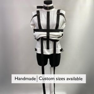 May include: A white garment with black straps and metal hardware, displayed on a mannequin. The garment has a high collar and is designed to secure the arms. The text "Handmade | Custom sizes available" is at the bottom.