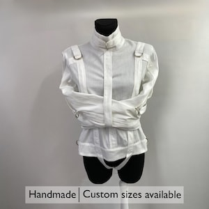 May include: White straight jacket with a high collar and straps, displayed on a mannequin. The jacket has metal rings and is designed to restrain the arms. The text "Handmade | Custom sizes available" is at the bottom.