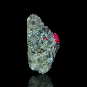 May include: A mineral specimen with a rough, textured surface. The mineral is a pale green colour with patches of red and dark flecks. The background is black, highlighting the mineral's details.