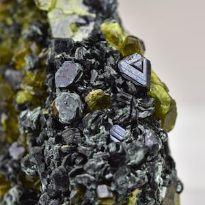 May include: Detailed image of a mineral specimen with dark, metallic crystals and bright, yellowish-green formations. A triangular, metallic crystal stands out amidst the cluster. The composition showcases the natural textures and colours of the mineral.