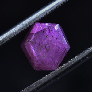 May include: A hexagonal, purple gemstone is held by tweezers against a black backdrop. The translucent gemstone has dark inclusions. The silver tweezers have a serrated edge. The gemstone is faceted.