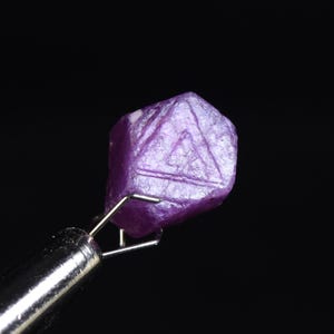 May include: A faceted, raw, purple gemstone held by a silver metal tweezer. The gemstone has a geometric shape with visible surface texture and a triangular pattern. The background is solid black.