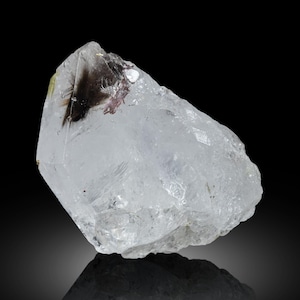 May include: A clear quartz crystal with dark inclusions and a slight purple hue. The crystal is irregularly shaped with a rough, textured surface. The image is set against a black background, highlighting the crystal's clarity and internal features.