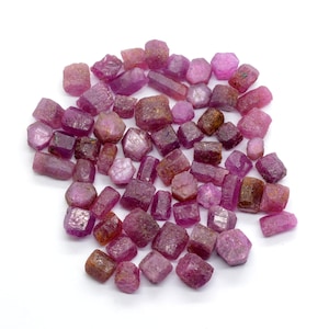 May include: A collection of pink ruby rough stones, each with a unique shape and size. The stones are arranged in a loose pile on a white background.