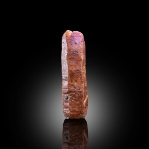 以下が含まれることがあります： 粗い質感の、細長いルビーの原石。深い赤褐色から鮮やかなピンク色へとグラデーションになっています。小さな黒いインクルージョンが全体に見られます。光沢のある黒い背景に、原石の反射が映っています。