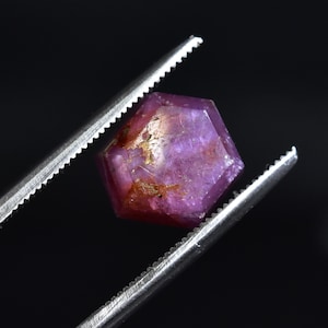 May include: A faceted, hexagonal ruby gemstone held by silver tweezers against a black backdrop. The ruby displays a deep pinkish-red hue with golden inclusions and a polished surface, showcasing its natural beauty.