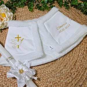 Peut inclure: Un ensemble en tissu blanc comprenant un tissu matelassé bordé de fausse fourrure, brodé d'une colombe et d'une croix dorées, avec le texte Clémence 29.10.2023. Une bougie blanche assortie porte un nœud en dentelle et un emblème doré.