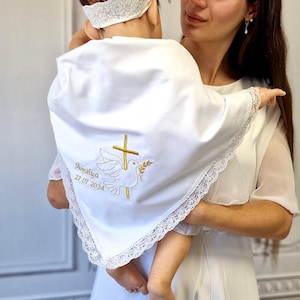 Può includere: Uno scialle battesimale triangolare bianco con un delicato bordo in pizzo. Il tessuto è ricamato con una croce dorata, una colomba bianca che tiene un ramoscello d'ulivo e il testo Amélya 27.07.2024. È visibile anche una fascia in pizzo bianco.