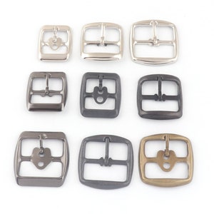 Könnte beinhalten: Neun Metall-Gürtelschnallen in verschiedenen Ausführungen, darunter Silber, Schwarz und Messing. Die Schnallen sind alle quadratisch mit einem Mittelbalken und zwei seitlichen Schlaufen.