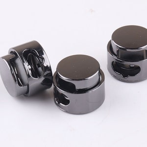 Könnte beinhalten: Drei schwarze Metall-Kordelstopper mit einem Federmechanismus. Die Stopper sind zum Sichern von Schnüren oder Bändern gedacht.