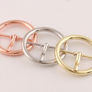 May include: Three round metal belt buckles in different finishes: rose gold, silver, and gold.