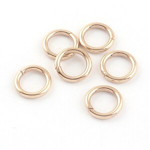 May include: Six gold-toned jump rings, also known as split rings, are arranged on a white surface. These rings are commonly used in jewellery making and crafting.
